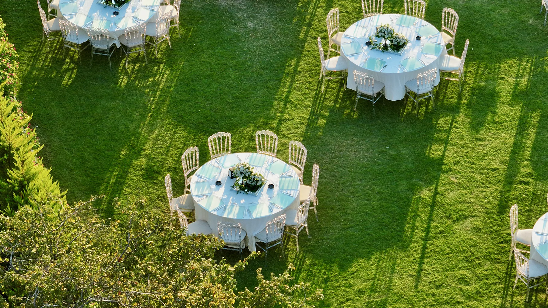 aerial-view-outdoor-event-tables-on-lawn-hmg-plus. Outdoor garden event with round tables, white linens, and clear chairs arranged on a green lawn for an upscale private gathering