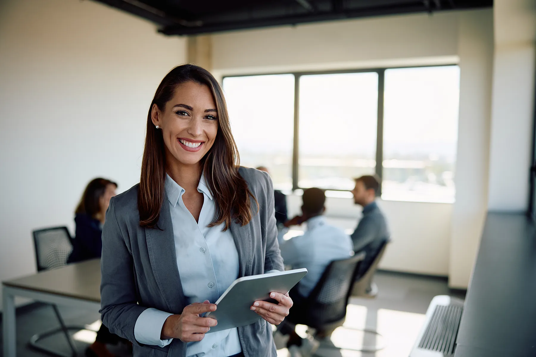 businesswoman-tablet-office-conference-room-team-meeting-hmg-plus. A smiling businesswoman in a gray blazer holding a tablet in a modern office conference room.
